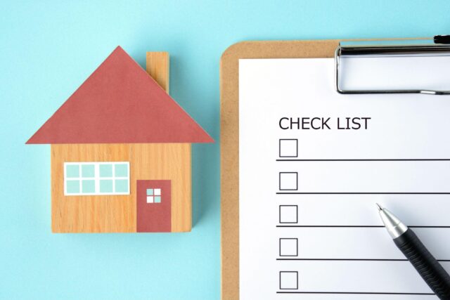 An image of a checklist board with a hand holding a pen, next to a cardboard house.